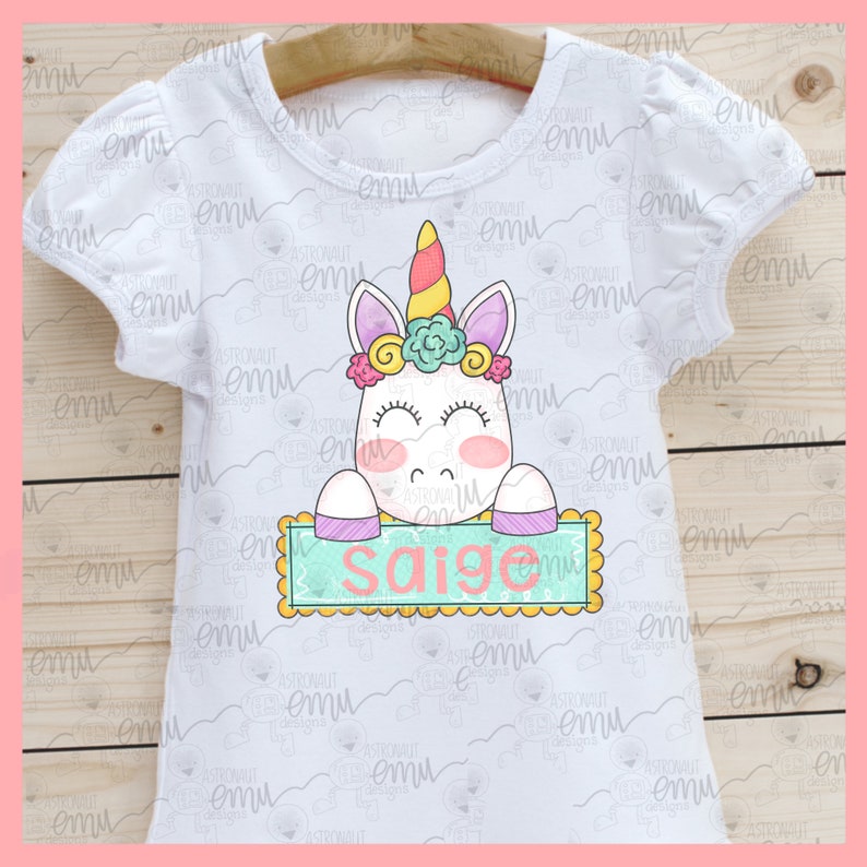 Op de afbeelding: Wit T-shirt met een eenhoorn afbeelding die een bord vasthoudt met de tekst "Saige". De eenhoorn heeft een regenbooghoorn, roze en paarse bloemen en gesloten ogen. Het bord is lichtblauw met een gele rand.