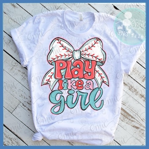 Op de afbeelding: Wit T-shirt met een honkbal-thema strik afbeelding en de tekst "Play like a girl" in rood, roze en teal. Het ontwerp is gecentreerd op de voorkant van het shirt.