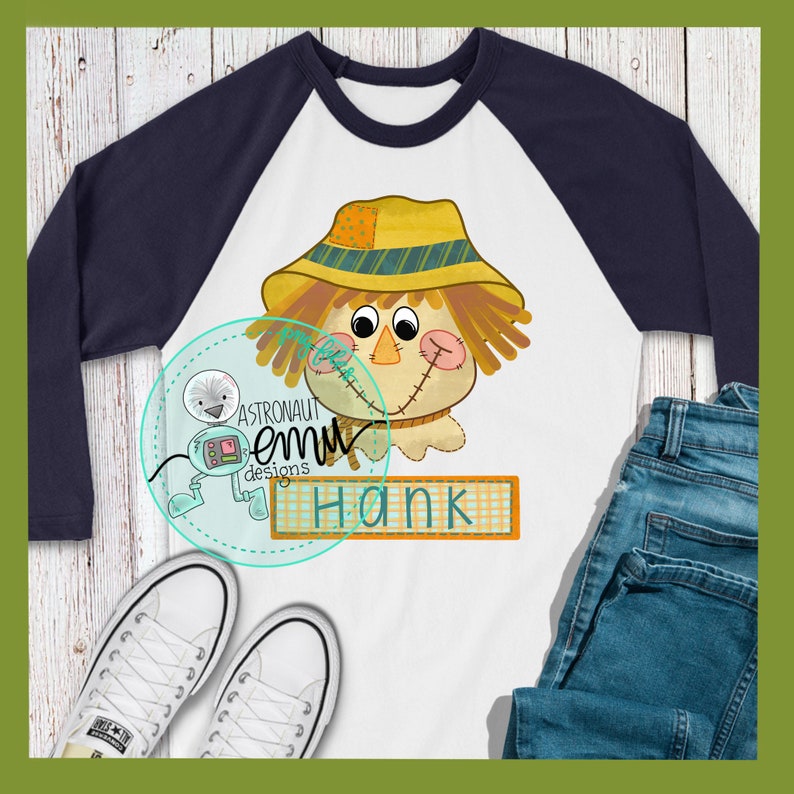 May include: A white and navy blue baseball tee shirt with a cartoon scarecrow wearing a yellow and brown hat. The shirt has the name "Hank" printed on a brown and white checkered banner.