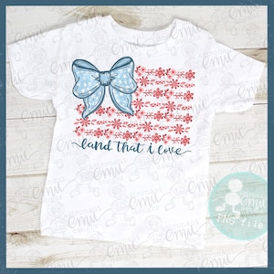 Pu&ograve; includere: T-shirt bianca con un design patriottico. Un fiocco azzurro con stelle bianche &egrave; in alto a sinistra, con una composizione floreale in rosso e rosa che ricorda una bandiera. Il testo "land that I love" &egrave; scritto in corsivo.