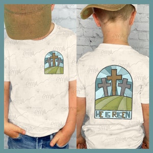 以下が含まれることがあります： 青い空と「HE IS RISEN」の文字が書かれた丘の上に3つの十字架が描かれた白いTシャツ。