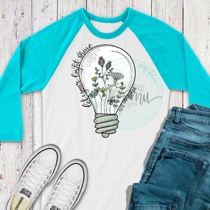 May include: A white long-sleeve baseball tee with a turquoise blue raglan sleeve. The shirt features a black and white illustration of a light bulb with flowers inside. The text "Let your light shine" is written in cursive on the side of the light bulb.