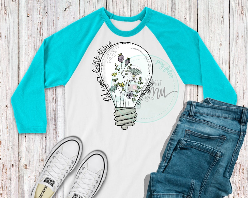 May include: A white long-sleeve baseball tee with a turquoise blue raglan sleeve. The shirt features a black and white illustration of a light bulb with flowers inside. The text "Let your light shine" is written in cursive on the side of the light bulb.