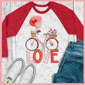 Pu&ograve; includere: Una maglietta bianca a maniche raglan con un design a maniche rosse. La maglietta presenta una bicicletta rosa con cuori e fiori rossi, e la parola "LOVE" in lettere rosa e rosse.