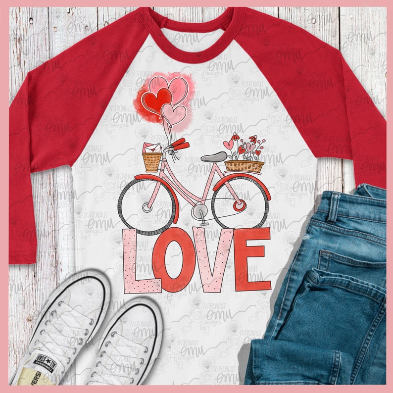 Pu&ograve; includere: Una maglietta bianca a maniche raglan con un design a maniche rosse. La maglietta presenta una bicicletta rosa con cuori e fiori rossi, e la parola "LOVE" in lettere rosa e rosse.