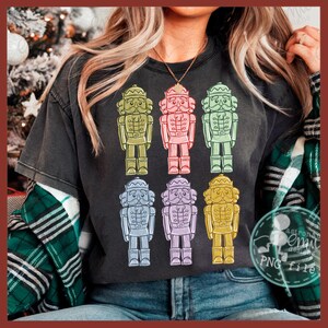 May include: Dark gray t-shirt featuring a colorful array of eight nutcracker soldier illustrations. The nutcrackers are in various colors, including green, pink, blue, and yellow. The design is a festive and whimsical representation of a holiday theme.