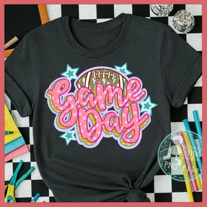 Puede incluir: Camiseta gris oscuro con un diseño colorido de "Game Day". El diseño incluye un balón de fútbol americano y estrellas, con las palabras en rosa, amarillo y azul. Una prenda de vestir informal.
