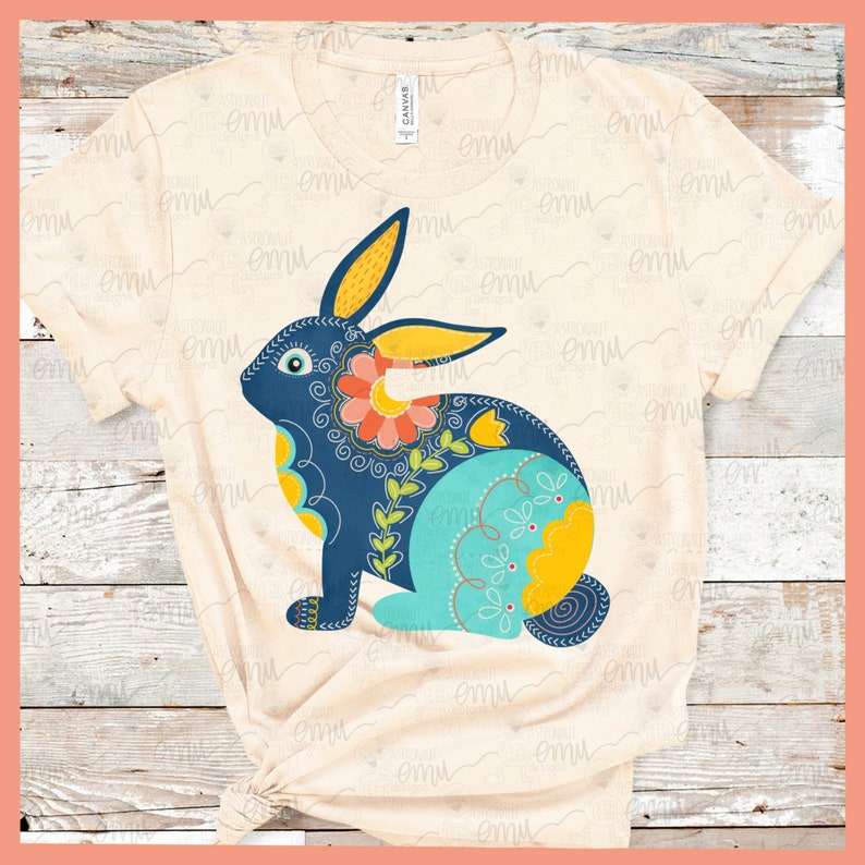 May include: A beige t-shirt with a blue, yellow, and green floral bunny design. The bunny is sitting on its hind legs with its ears up.