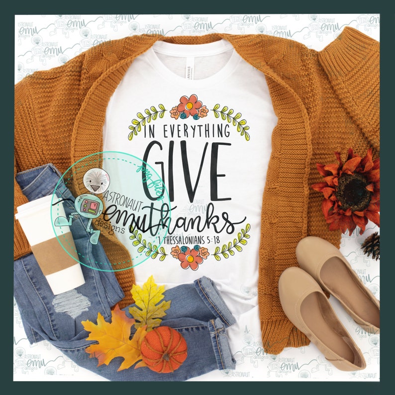 May include: A white t-shirt with a floral design and the text "In everything give thanks 1 Thessalonians 5:18". The shirt is styled with a brown cardigan, blue jeans, a coffee cup, and fall-themed accessories.