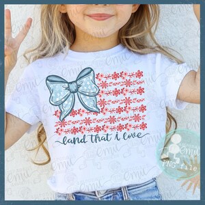 Pu&ograve; includere: T-shirt bianca con fiocco azzurro chiaro con motivo a stelle e un design floreale rosso e rosa. Il testo "land that I love" &egrave; scritto in corsivo. Una maglietta a tema patriottico.