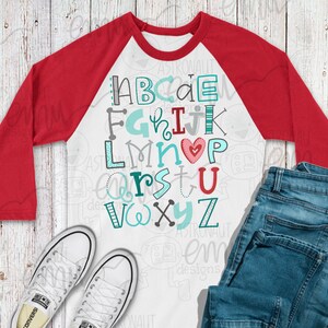May include: A white and red baseball tee shirt with a colorful alphabet design. The letters are in different fonts and colors, with a red heart in the center. The shirt has a white front and red sleeves.