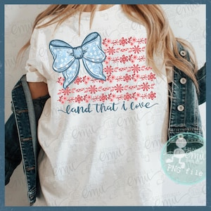 Pu&ograve; includere: T-shirt bianca con un fiocco blu con motivo a stelle e un disegno floreale in rosso e rosa, che ricorda una bandiera. Il testo "land that I love" &egrave; scritto in corsivo sotto.