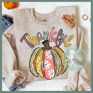 Może przedstawiać: Beżowa bluza z kolorową grafiką przedstawiającą dynię i napis "Thankful" w fantazyjnym kroju pisma. Dynia jest ozdobiona kwiatami i liśćmi.