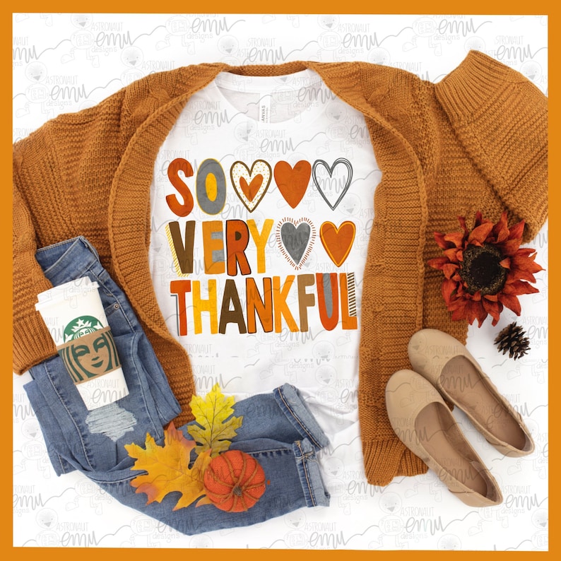 May include: A white t-shirt with the text "SO VERY THANKFUL" in colorful letters and hearts. The shirt is styled with a brown cardigan, blue jeans, a coffee cup, fall leaves, and a small pumpkin.