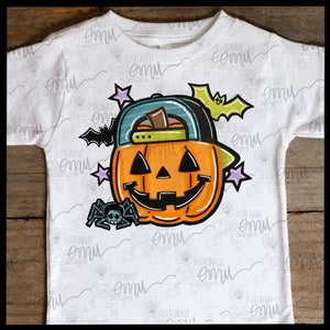 May include: A white t-shirt with a cartoon illustration of a jack-o'-lantern wearing a green and blue baseball cap. The jack-o'-lantern has a friendly face and is surrounded by stars, a bat, and a spider.