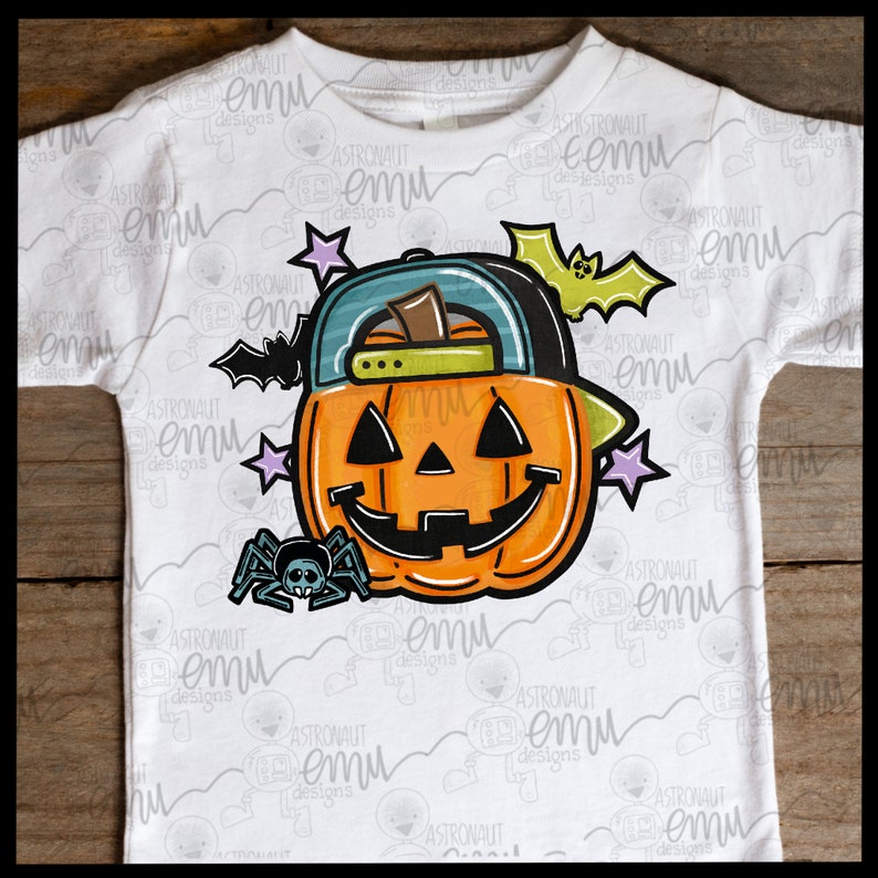 May include: A white t-shirt with a cartoon illustration of a jack-o'-lantern wearing a green and blue baseball cap. The jack-o'-lantern has a friendly face and is surrounded by stars, a bat, and a spider.