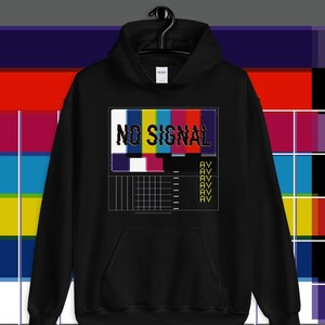 May include: Black hoodie featuring a "NO SIGNAL" design. The graphic shows a colourful test pattern with the words "NO SIGNAL" in a distressed font. The design includes the letters "AV" repeated below the test pattern.