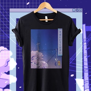 May include: Black t-shirt featuring a graphic of a cityscape at night with cherry blossoms and the Japanese text "そばにいてください" which translates to "Please be by my side".
