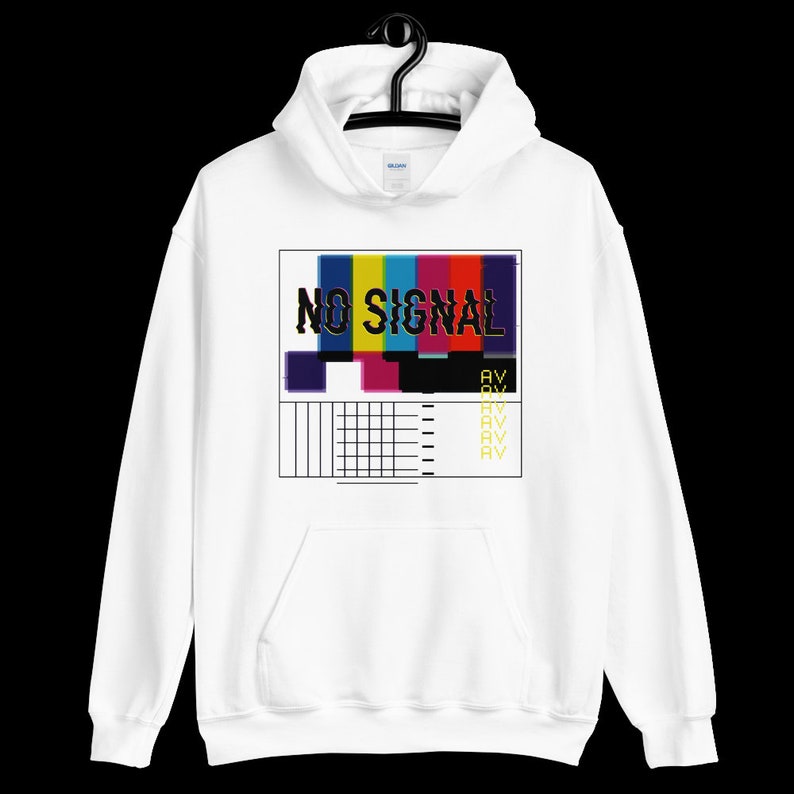 May include: White hoodie featuring a graphic that reads "NO SIGNAL." The design includes a colourful test pattern with blue, yellow, pink, and red bars, alongside black and white grid lines. The sweatshirt has a front pocket and a drawstring hood.