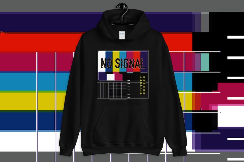 May include: Black hoodie featuring a "NO SIGNAL" design. The graphic shows a colourful test pattern with the words "NO SIGNAL" in a distressed font. The design includes the letters "AV" repeated below the test pattern.