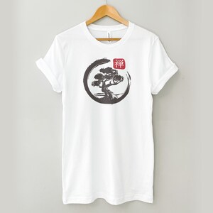 Puede incluir: Camiseta blanca con una ilustraci&oacute;n en blanco y negro de un &aacute;rbol en un c&iacute;rculo con la palabra japonesa "Zen" en rojo.