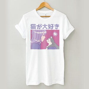 Puede incluir: Camiseta blanca con una ilustraci&oacute;n rosa y morada de una chica con cabello morado mirando a un gato. El texto "猫が好き" est&aacute; impreso en morado en la camiseta.