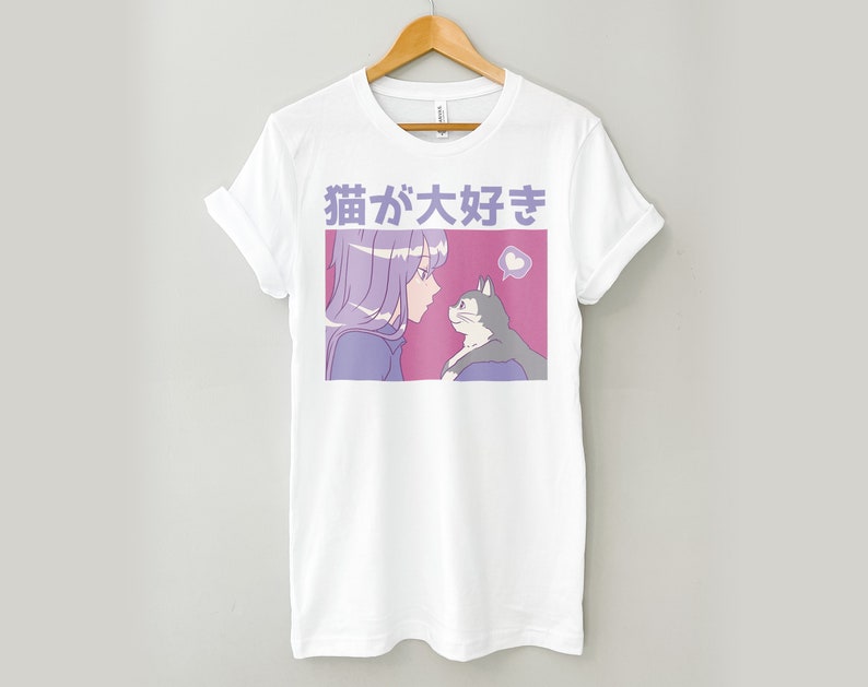 Puede incluir: Camiseta blanca con una ilustraci&oacute;n rosa y morada de una chica con cabello morado mirando a un gato. El texto "猫が好き" est&aacute; impreso en morado en la camiseta.