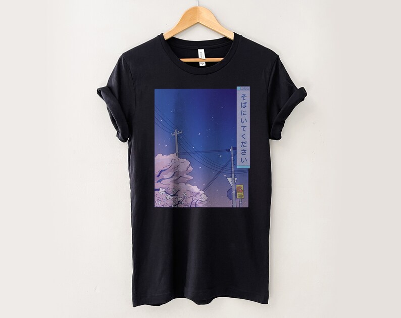 May include: Black t-shirt featuring a graphic print of a cityscape at night with a pink cherry blossom tree and the Japanese text "そばにいってほしい" which translates to "I want to be by your side".