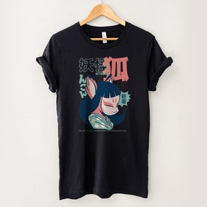 Peut inclure: T-shirt noir avec un imprim&eacute; graphique d'un kitsune (esprit de renard japonais) de dessin anim&eacute; avec un visage rose et des v&ecirc;tements bleus et verts. Le texte "Kitsune" et des caract&egrave;res japonais sont imprim&eacute;s sur le t-shirt.