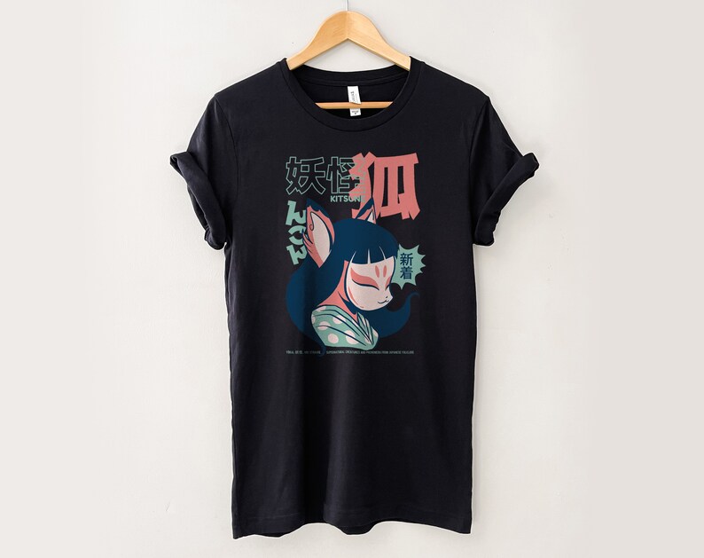 Peut inclure: T-shirt noir avec un imprim&eacute; graphique d'un kitsune (esprit de renard japonais) de dessin anim&eacute; avec un visage rose et des v&ecirc;tements bleus et verts. Le texte "Kitsune" et des caract&egrave;res japonais sont imprim&eacute;s sur le t-shirt.