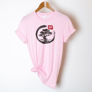 Puede incluir: Camiseta rosa con un gr&aacute;fico en blanco y negro de un &aacute;rbol en un c&iacute;rculo con la palabra japonesa "Zen" en rojo.