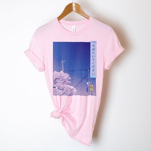 May include: Pink t-shirt featuring a graphic print of a cityscape at night with cherry blossoms and the Japanese text "そこにいてください" which translates to "Stay there".