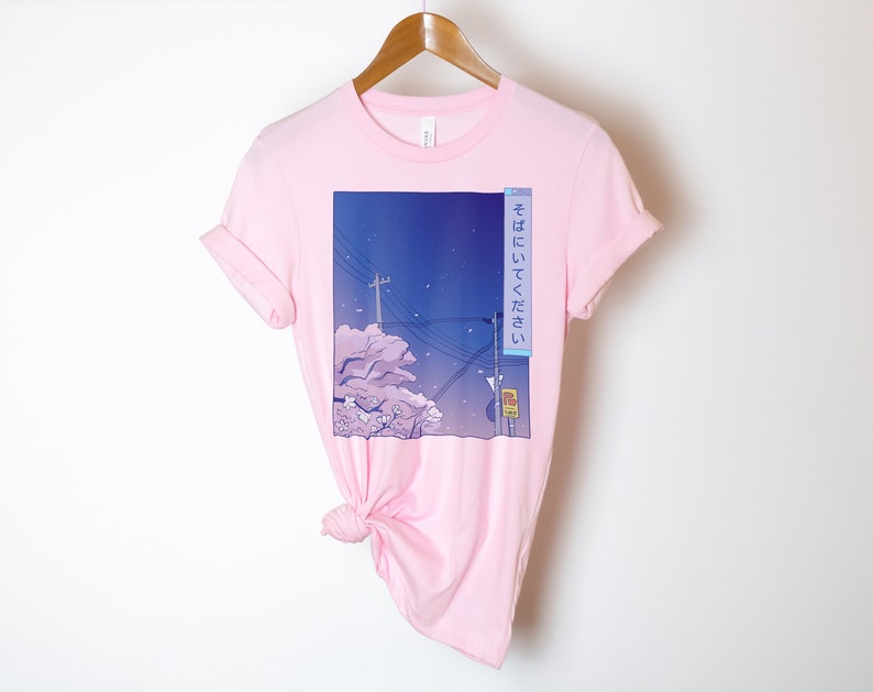 May include: Pink t-shirt featuring a graphic print of a cityscape at night with cherry blossoms and the Japanese text "そこにいてください" which translates to "Stay there".