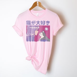 Puede incluir: Camiseta rosa con un estampado gr&aacute;fico de una chica con el pelo morado mirando a un gato. El texto "猫が好き" est&aacute; impreso en la camiseta.