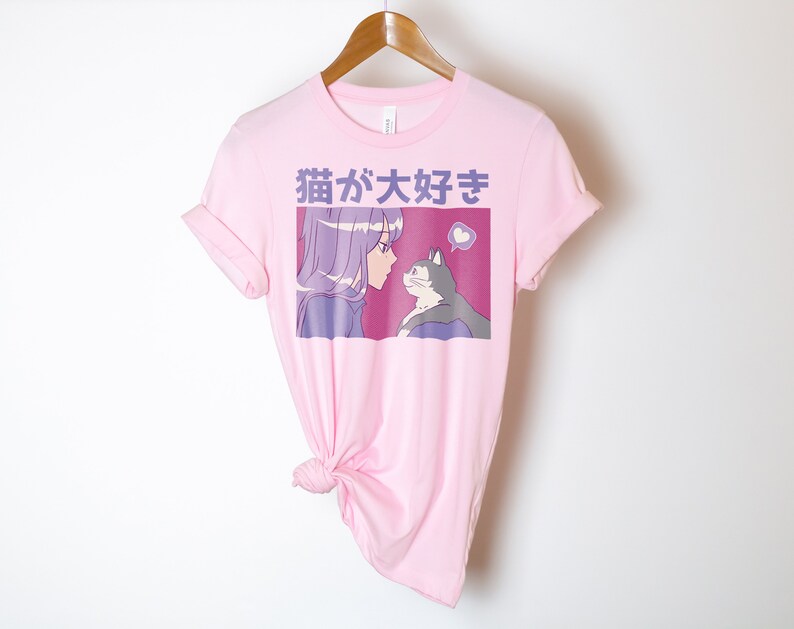 Puede incluir: Camiseta rosa con un estampado gr&aacute;fico de una chica con el pelo morado mirando a un gato. El texto "猫が好き" est&aacute; impreso en la camiseta.