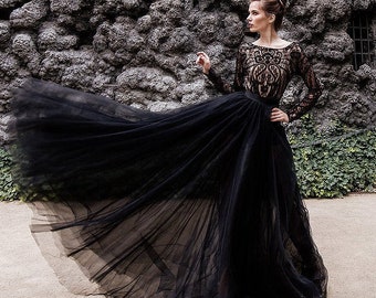 May include: A woman in a long black gown with a flowing skirt stands in a garden. The dress features a detailed lace bodice and long sleeves. The background includes a stone wall, a statue, and white flowers.