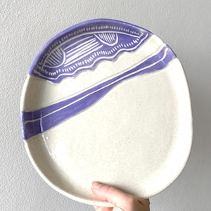 May include: Oval ceramic plate with a speckled off-white surface. The plate features a decorative border and a diagonal stripe in a lavender colour. The plate is held by a hand against a light grey background.