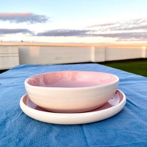 Puede incluir: Un cuenco de cerámica rosa claro sobre un plato de cerámica rosa claro a juego. El cuenco tiene un interior liso y redondeado con un borde ligeramente grueso, mientras que el plato presenta una base plana y un borde elevado, ambos brillantes.