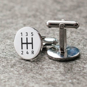 Könnte beinhalten: Ein Paar silberne Manschettenknöpfe mit einem weißen runden Design, das ein Schaltmuster mit den Zahlen 1, 2, 3, 4, 5 und R zeigt.