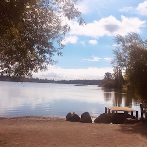 May include: A view of a lake with a wooden platform on the shore. The lake is calm and the water is reflecting the blue sky. There are trees on the shore and a few large rocks on the beach.