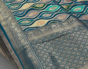 May include: A detailed shot of a teal and silver textile with an intricate pattern. The fabric features a repeating wave-like design in teal, gray, and gold, with a decorative border. The textile is likely used for clothing or home decor.