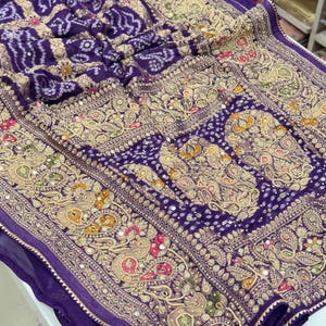 Pu&ograve; includere: Primo piano di un tessuto viola con ricami dorati intricati. Il design presenta motivi paisley, motivi floreali e specchi. Il tessuto ha un aspetto strutturato, con una tonalit&agrave; viola intenso e dettagli dorati.