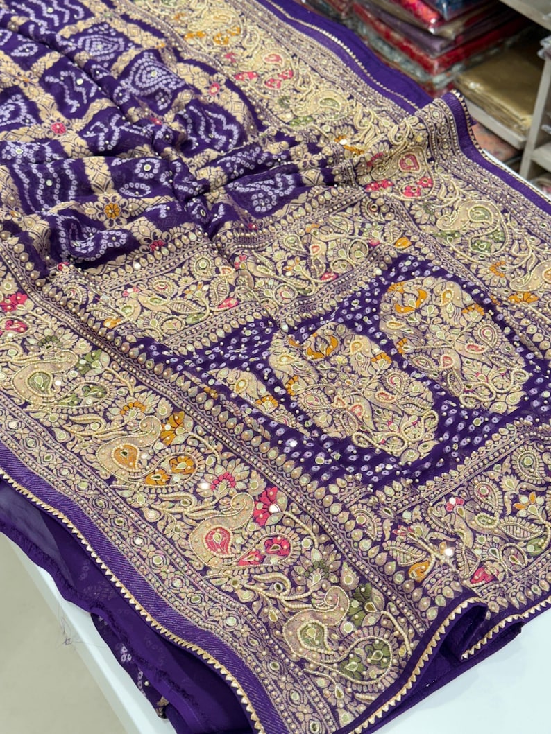 Pu&ograve; includere: Primo piano di un tessuto viola con ricami dorati intricati. Il design presenta motivi paisley, motivi floreali e specchi. Il tessuto ha un aspetto strutturato, con una tonalit&agrave; viola intenso e dettagli dorati.