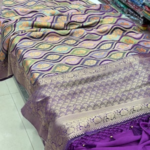 May include: A purple and gold patterned silk saree with a purple border and gold trim. The saree has a woven design and is draped over a white surface.