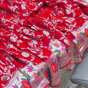 Puede incluir: Un textil rojo vibrante con un detallado patr&oacute;n floral plateado. La tela est&aacute; drapeada, mostrando el intrincado dise&ntilde;o. El borde presenta un ribete plateado con acentos florales coloridos, agregando un toque de elegancia.