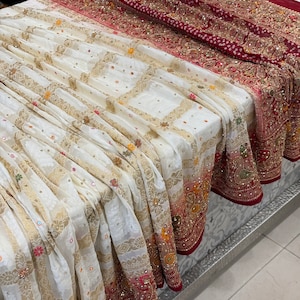 May include: A white and red embroidered sari with gold accents. The sari is folded and draped on a table.