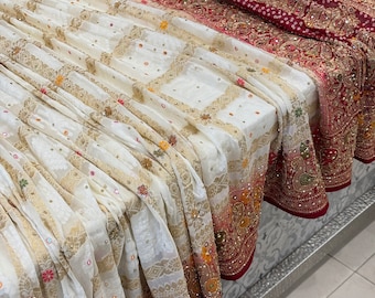 Puede incluir: Un sari blanco y rojo bordado con detalles dorados. El sari está doblado y drapeado sobre una mesa.