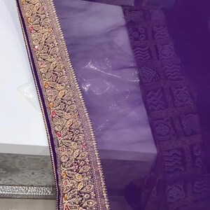 Pu&ograve; includere: Primo piano di un tessuto viola con un bordo ricamato ornato. Il bordo presenta disegni floreali e paisley dorati, rossi e verdi. Il tessuto sembra essere un materiale trasparente, forse una sciarpa o uno scialle.