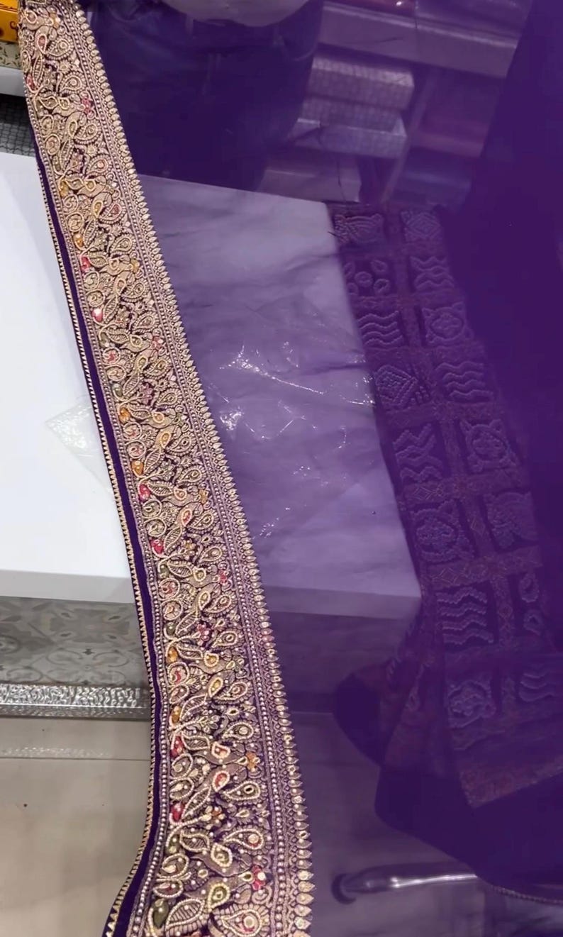Pu&ograve; includere: Primo piano di un tessuto viola con un bordo ricamato ornato. Il bordo presenta disegni floreali e paisley dorati, rossi e verdi. Il tessuto sembra essere un materiale trasparente, forse una sciarpa o uno scialle.