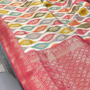 May include: A white and grey patterned saree with a pink and gold border. The saree has a wavy design with colorful accents.